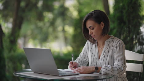 Student-Woman-Brunette-arabic-Hispanic-ethnos-studying-remotely-via-the-Internet-while-sitting-in-a-summer-cafe-with-a-laptop-and-writing-down-a-pen-and-a-notebook-while-taking-notes-on-the-webinar