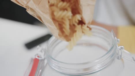 close up of pasta pouring from brown paper bag into storage jar in kitchen, in slow motion