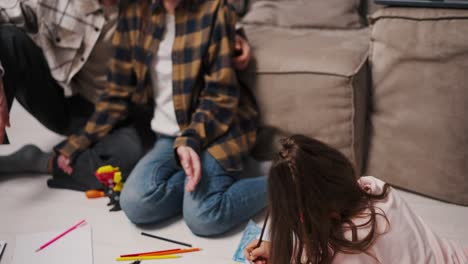 cerca, una pequeña chica morena con ropa rosa dibuja tendida en el suelo sus padres la miran un hombre moreno con paja en una camisa a cuadros y su esposa morena en una camiseta blanca y camisa a cuadrados cerca de un sofá gris y marrón en un apartamento estudio moderno