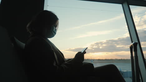 silhouette of a woman in a protective mask, sitting at a large window in the airport terminal, using a smartphone