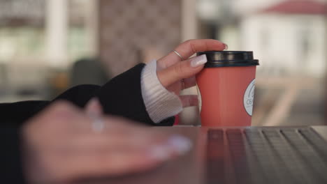 close-up of lady's hand gently turning coffee cup near laptop, focusing on manicured fingers and rings against laptop keyboard, with blurred background of chair