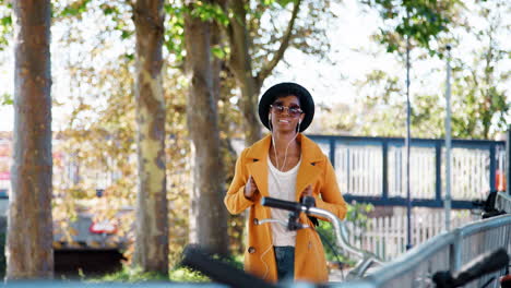 Fashionable-young-black-woman-wearing-a-hat,-sunglasses,-blue-jeans-and-a-yellow-pea-coat-walking-towards-camera-along-a-treelined-street-on-a-sunny-day-listening-to-music-with-earphones,-selective-focus