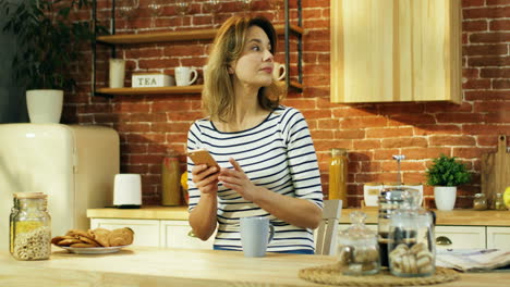 Mujer-Joven-Y-Atractiva-Sentada-En-La-Cocina-Con-Un-Café-Por-La-Mañana-Y-Tocando-O-Escribiendo-En-El-Teléfono-Inteligente