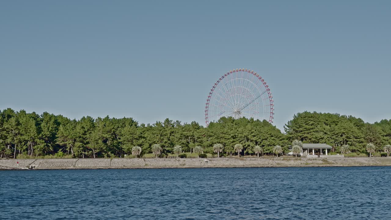 A wide, bright shot across water toward a green, tree-lined park with a large white Ferris wheel rising high above the foliage