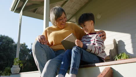 Feliz-Abuela-Birracial-Y-Nieto-Usando-Tableta-Sentada-En-La-Terraza,-Cámara-Lenta