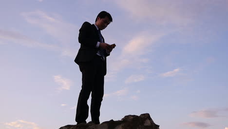 businessman using smartphone on a mountaintop at sunset