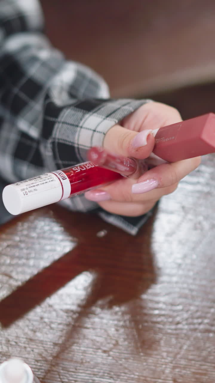 primo piano di mani che testano campioni lucidi di rossetto sulle dita, donna che apre il tubetto e preme l'applicatore, tavolo riflettente e giochi d'ombra, movimento attento che mostra la texture e la lucentezza, atmosfera da dimostrazione informale del prodotto