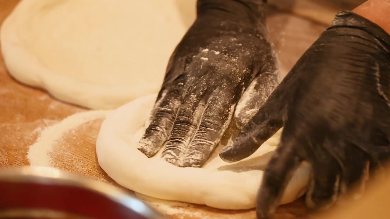 Primo piano di mani con guanti neri che stendono impasto fresco per pizza su una superficie di legno infarinata