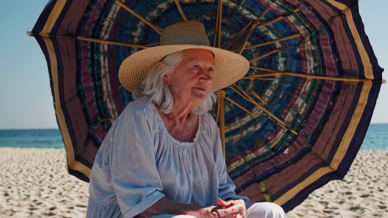 Senior woman wearing straw hat sitting under colorful beach umbrella on sandy shore, enjoying peaceful moment by the sea during summer vacation