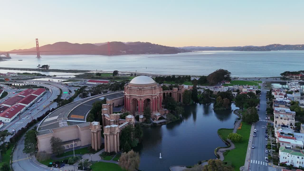 Drone footage of the Palace of Fine Arts in San Francisco with the Golden Gate Bridge in the distance, captured from a high aerial perspective at sunset
