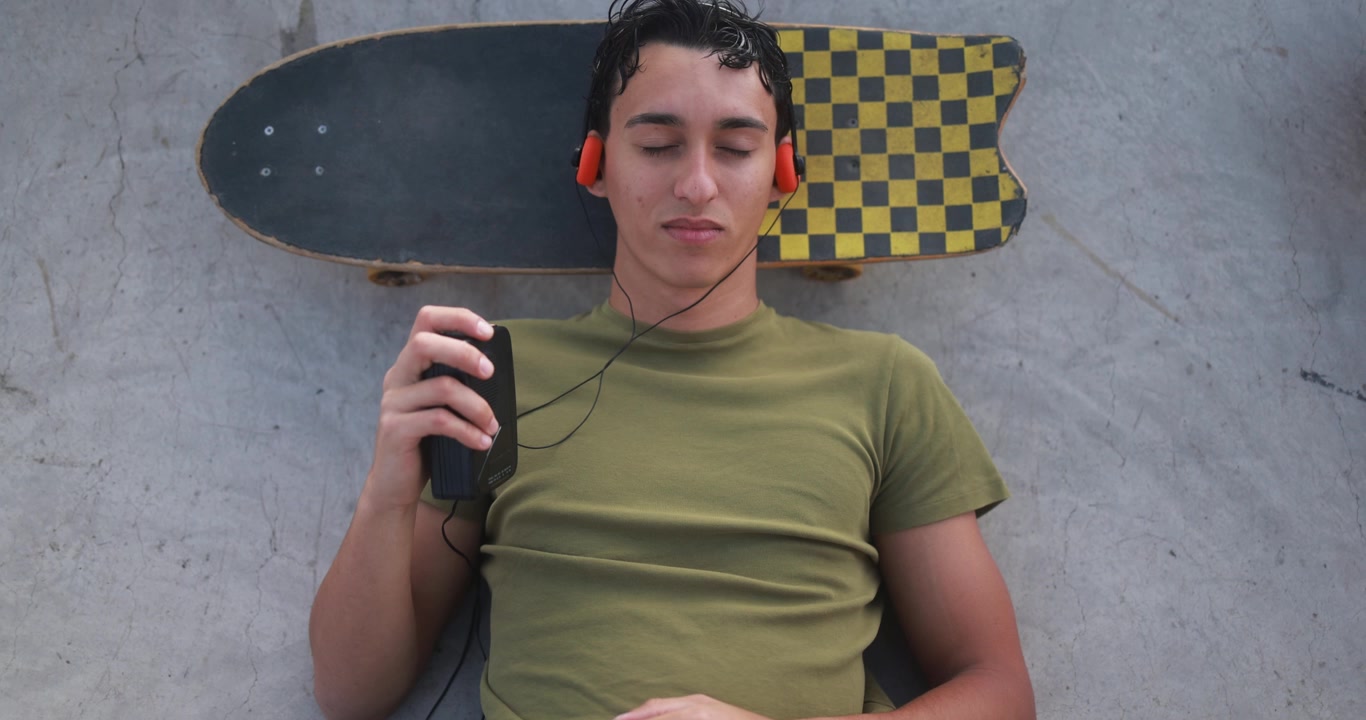 Giovane sdraiato sul cemento con lo skateboard, che ascolta musica con un lettore di cassette