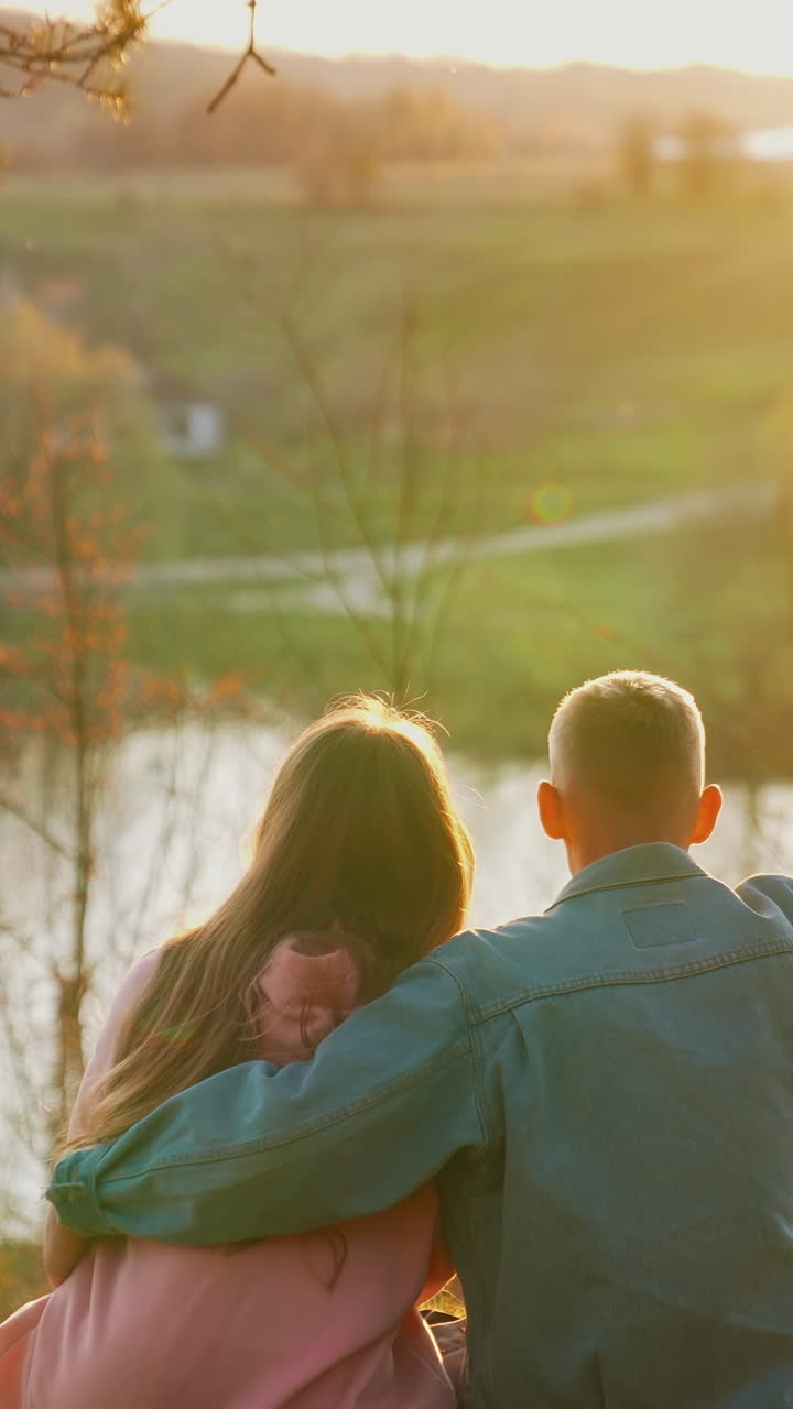 Young people sitting on grass and looking at sunset near the river. Guy embracing his girlfriend against autumn landscape in the evening. Vertical video