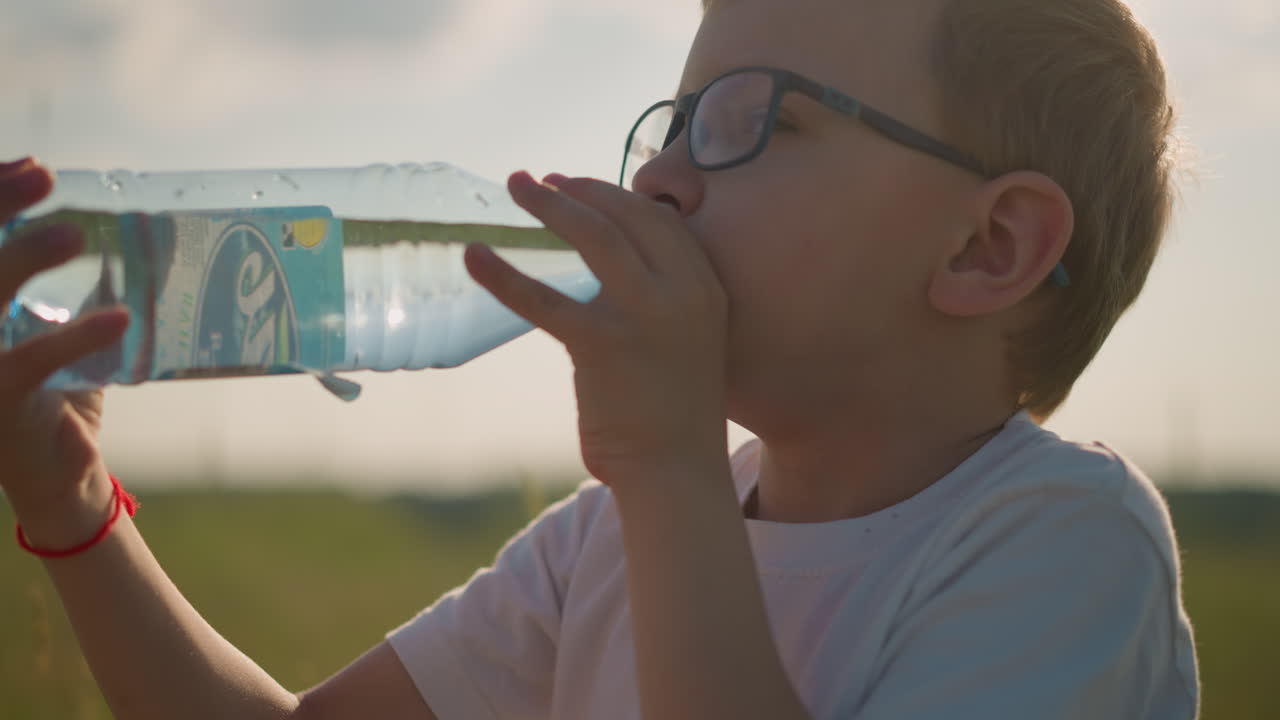 A young boy wearing a white top and glasses, with a red band on his wrist, is sitting in a grassy field, drinking water from a clear plastic bottle. moment of hydration in a natural, outdoor setting