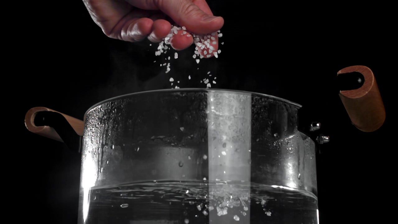 Hand Sprinkling Coarse Salt into Boiling Water in a Glass Pot