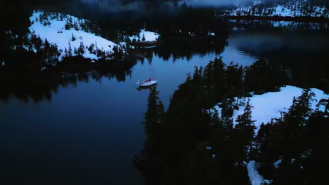 Aerial-view-of-a-sailboat,-moving-through-snowy-island-narrows,-in-gloomy-Alaska