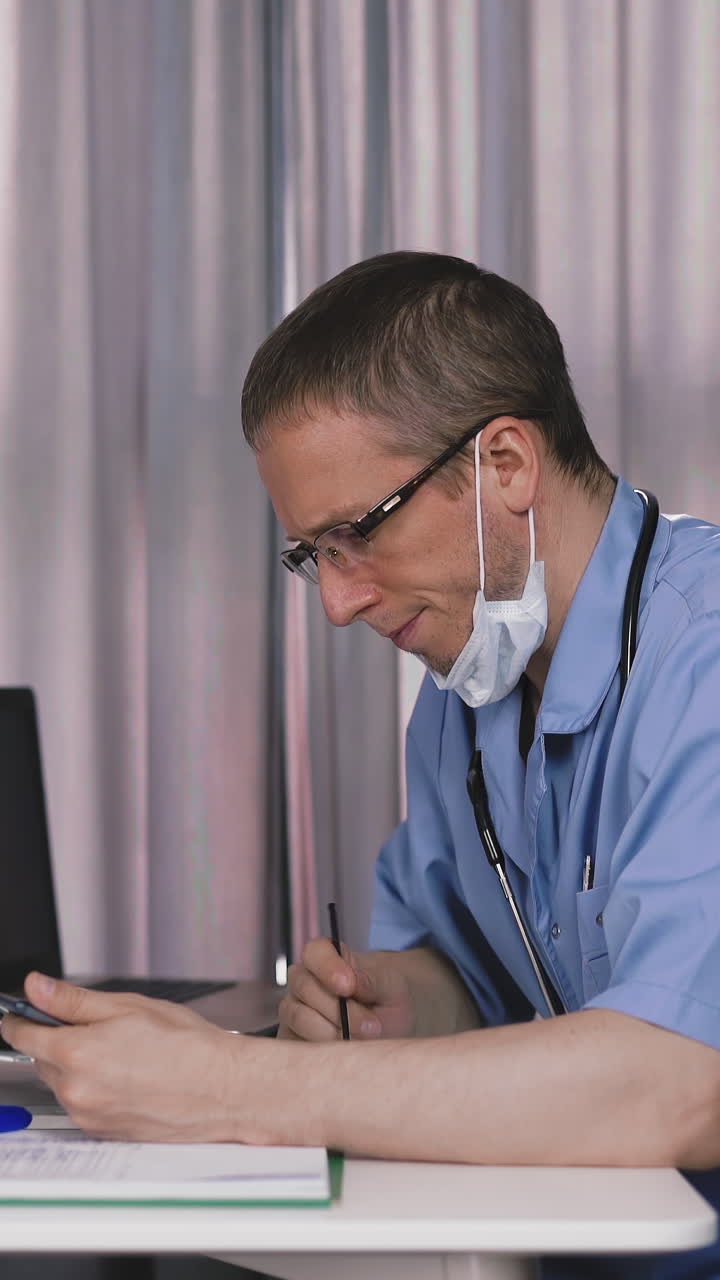 young bearded doctor in protective mask and glasses, sits in his office at table, holds stylus, online consultation with patient, smiles