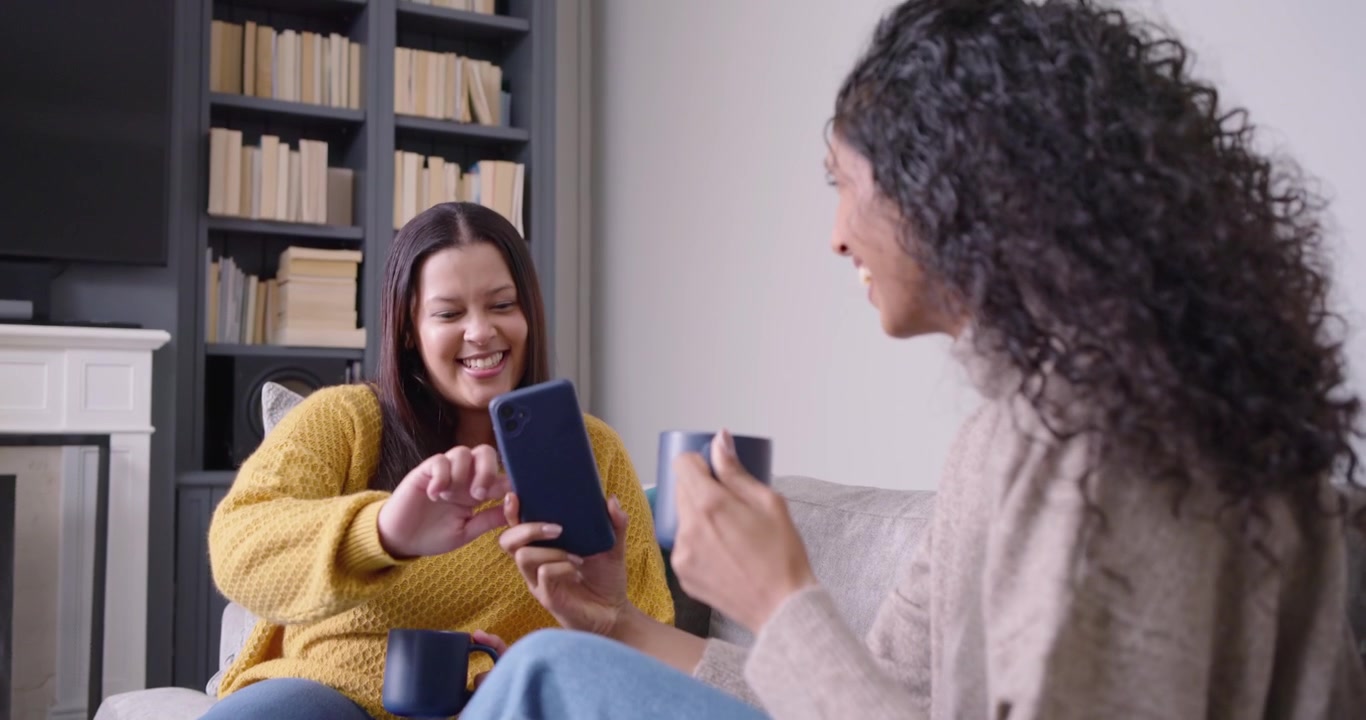 Due amiche sul divano, quella con il maglione beige mostra il telefono, quella con il maglione giallo tocca lo schermo, ridendo con le tazze.