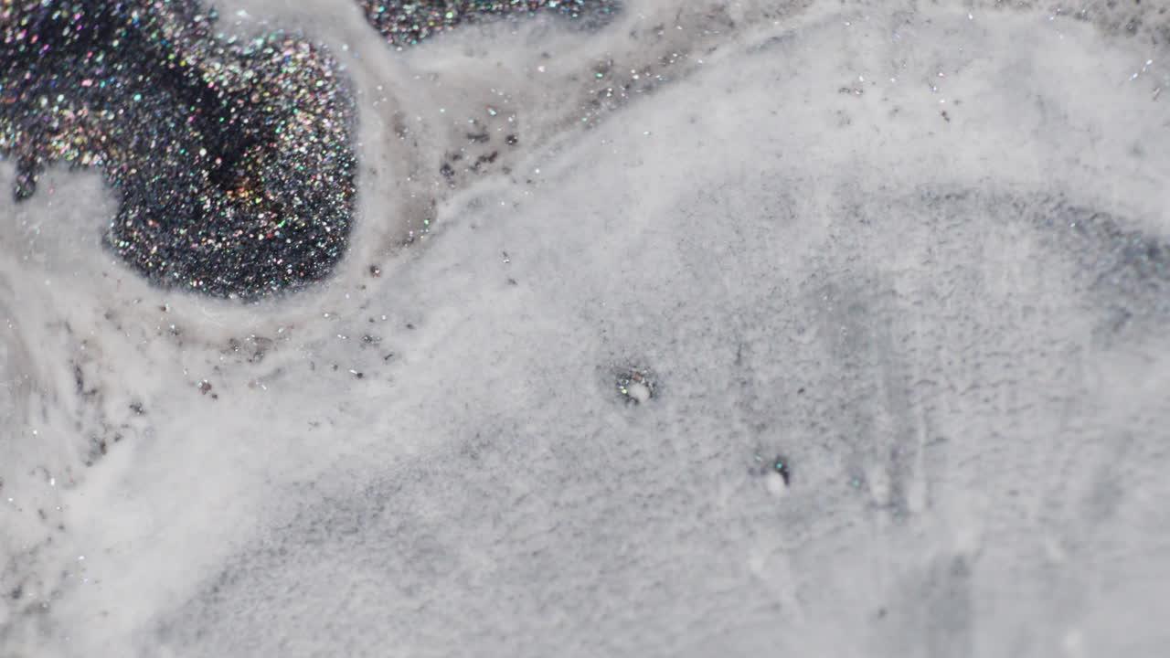 A milky fluid slowly spreads across glittery black and grey ink in a macro shot, eventually covering most of the shimmering surface