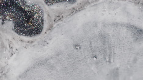 a milky fluid slowly spreads across glittery black and grey ink in a macro shot, eventually covering most of the shimmering surface