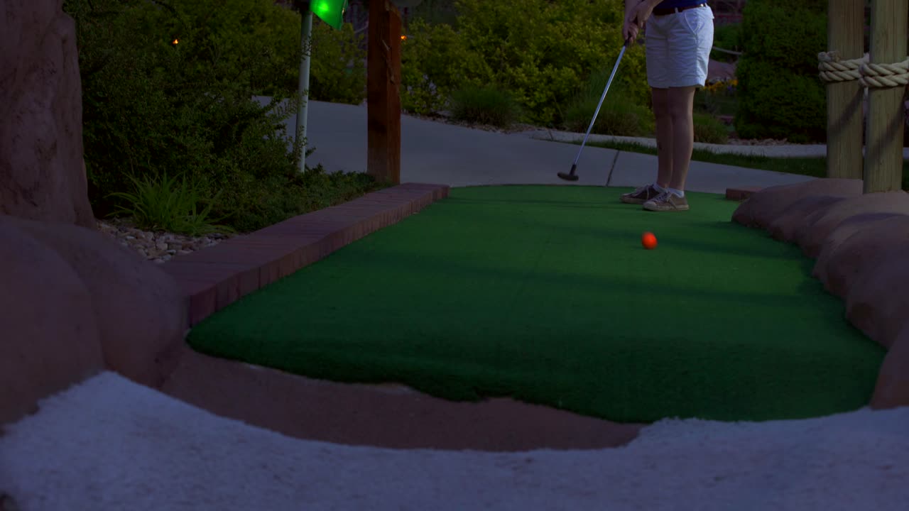 A caucasian girl mini golfing an orange golf ball hitting it towards the camera