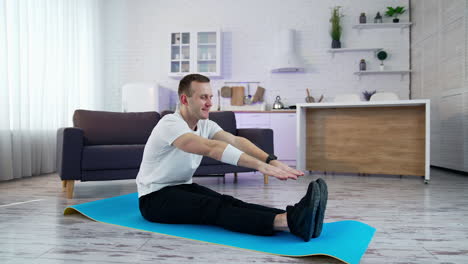 young man exercising at home. man sitting on a mat and trying to touch his feet on kitchen background. difficulties of physical training during lockdown period.