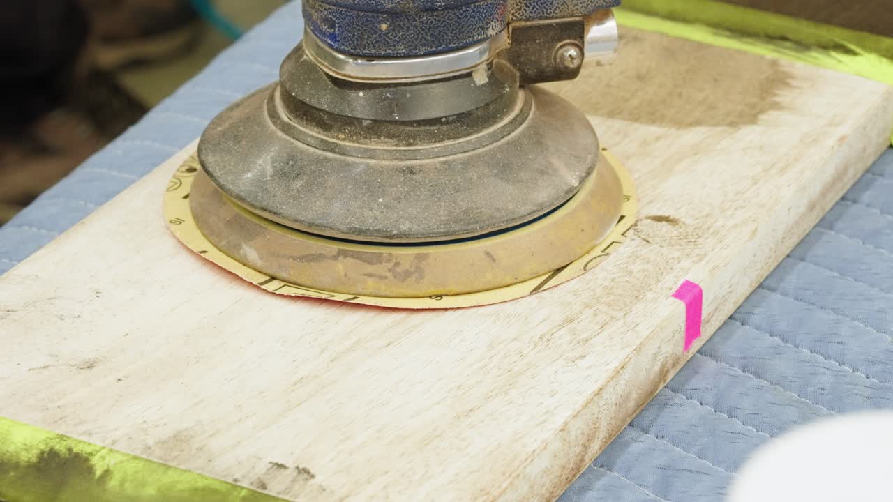 Orbital sander rests on bench as out-of-focus person walks around workshop