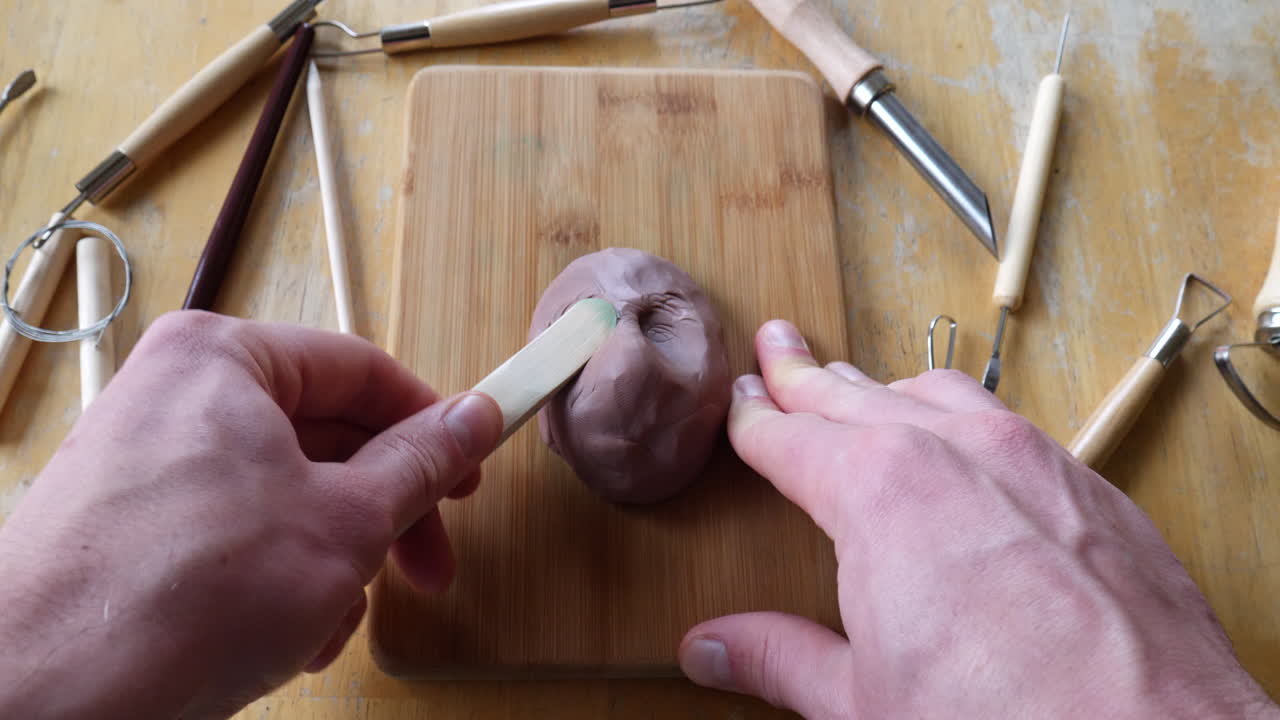 An artist sculpting brown modeling clay with tools to make a face mask figurine in his art studio