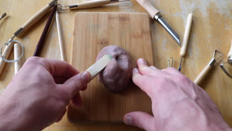 an artist sculpting brown modeling clay with tools to make a face mask figurine in his art studio