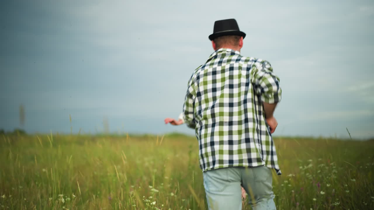 검은 모자와 체크 셔츠를 입은 남자가 하 드레스와 모자를 입은 여자를 따라 구름이 은 하늘 아래 잔디을 가로질러 달린다.