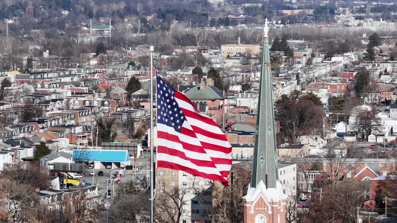 Bandiera americana che sventola su un quartiere e una chiesa nella città di York, USA. Giornata di sole con alberi spogli su una collina in città. Orbita aerea ravvicinata. Stagione invernale.