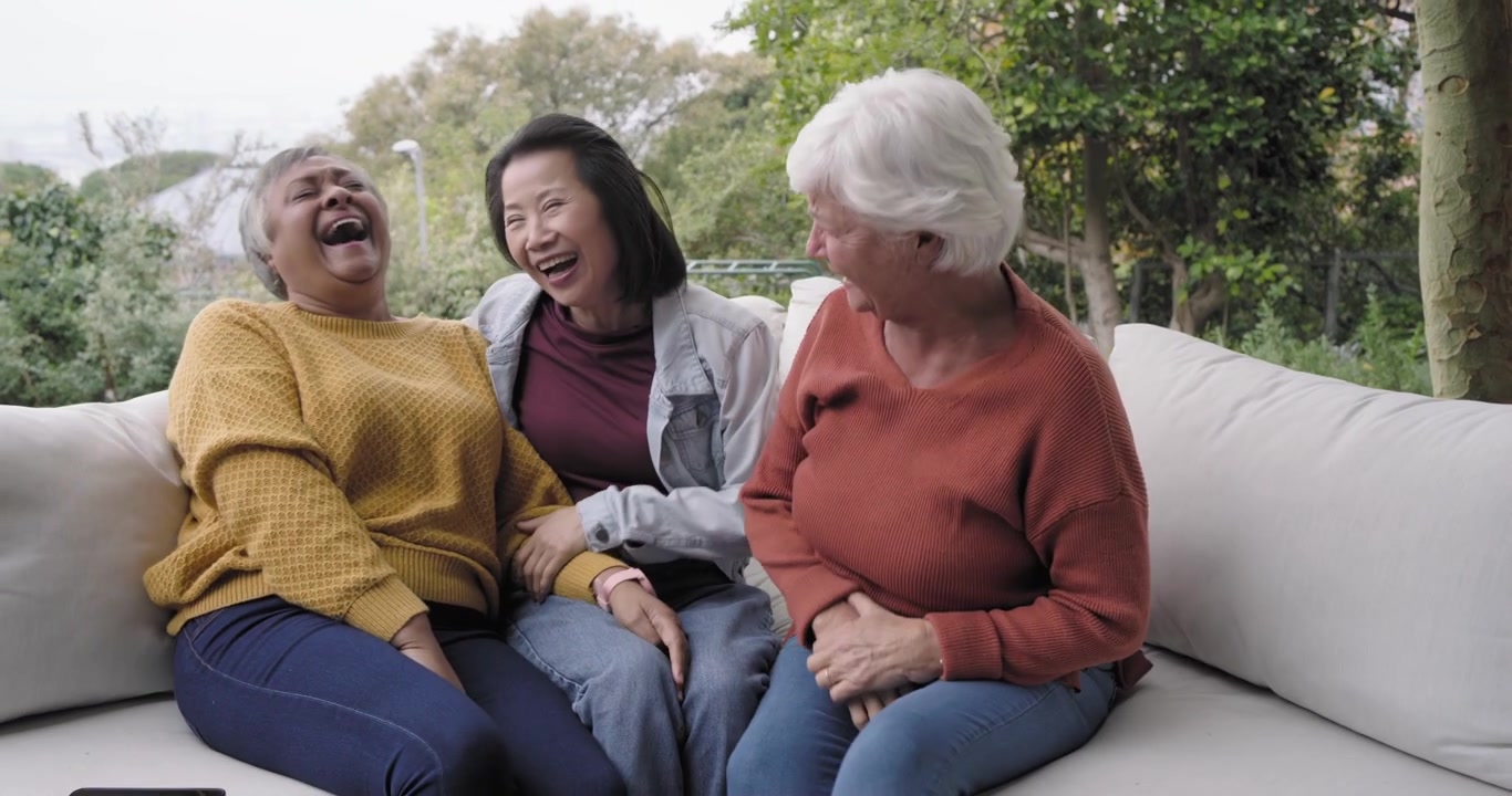 Diverse female friends in sweaters laughing at inside joke, leaning on garden sofa with phone