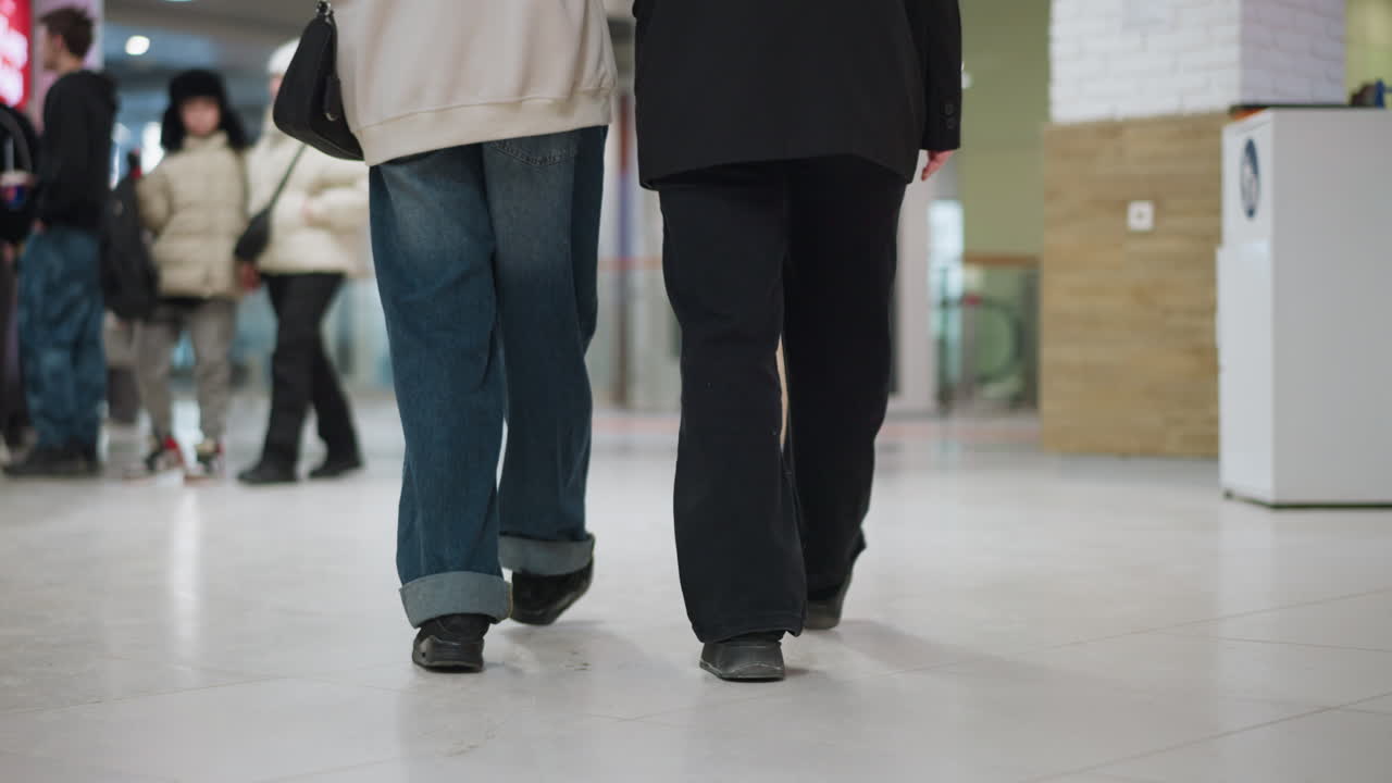Lower angle view of two ladies walking through bright modern mall corridor, casual outfits and relaxed pace, interacting while passing people and shops, representing urban lifestyle
