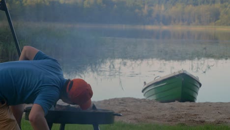 man kneeling, blowing firewoods and charcoal burning on a metal griller to cook barbeque at the lake shore in poland. - wide shot