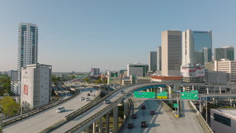 Forwards-fly-above-large-multilevel-and-multilane-highway-interchange-in-city.-Heavy-traffic-between-high-rise-buildings.-Miami,-USA