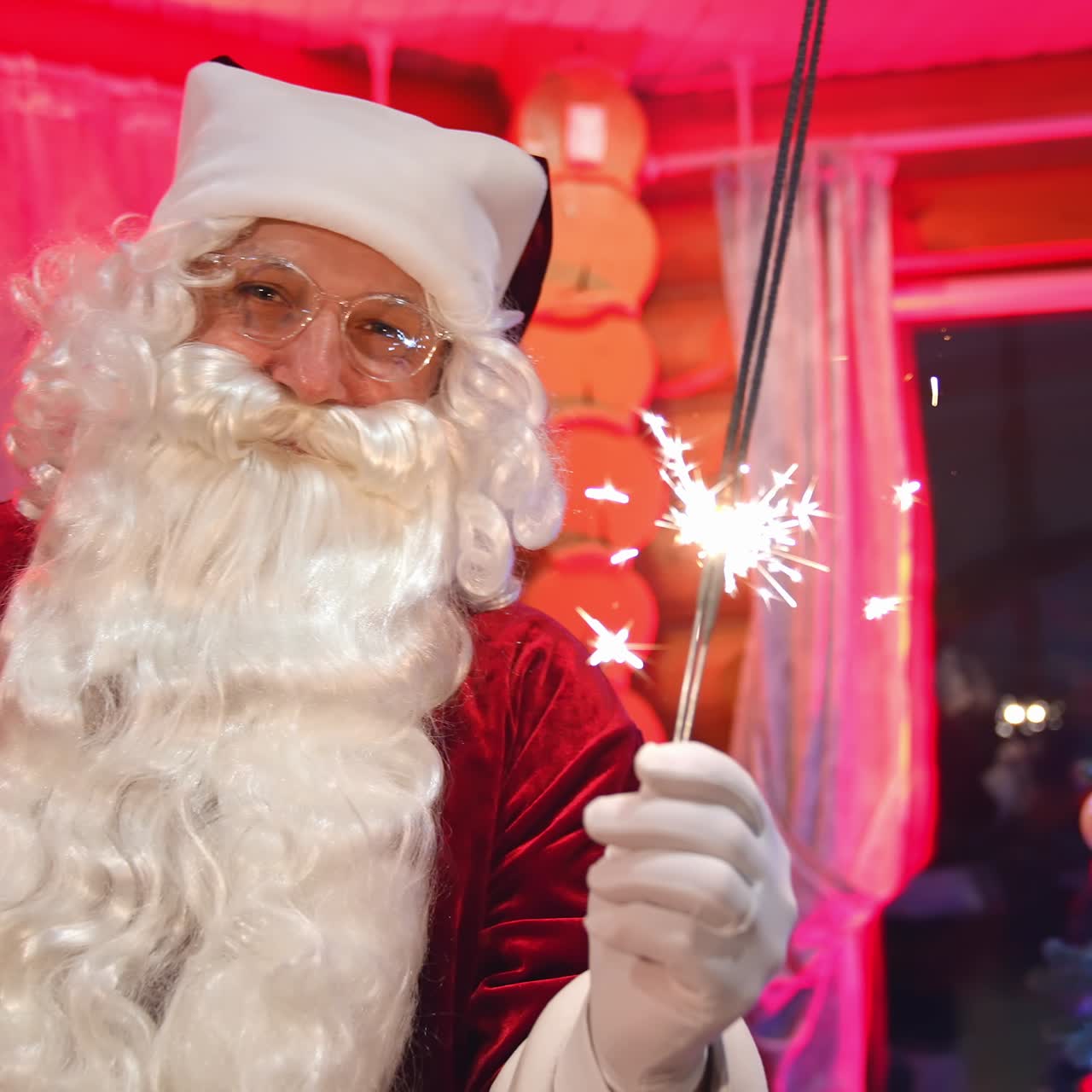 Portrait of Santa Claus with sparkler lights. Happy Santa with white beard wearing glasses standing on Christmas tree backdrop and looking on camera
