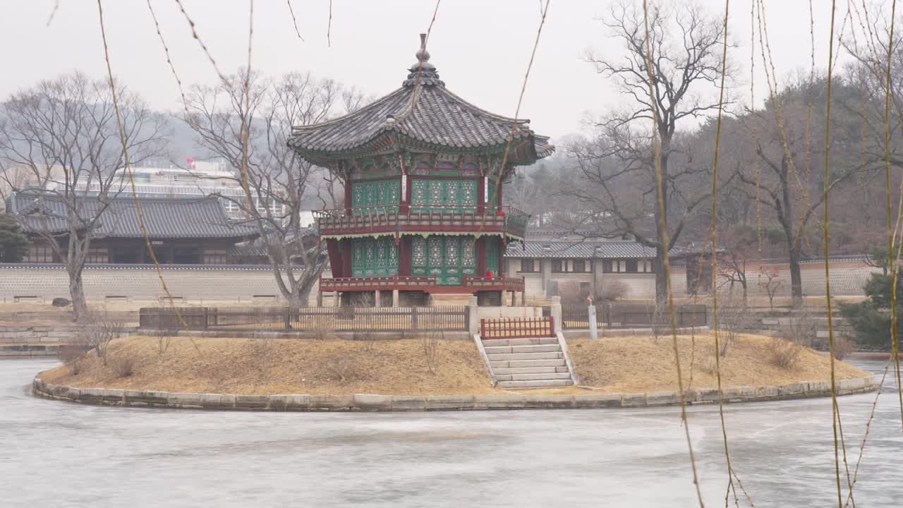 Paesaggio in slow motion dell'edificio del tempio-padiglione Hyangwonjeong su un'isola circondata da un lago ghiacciato e alberi con canne in primo piano, nei giardini del Palazzo Gyeongbokgung a Seoul, Corea del Sud. Viaggio in Asia.