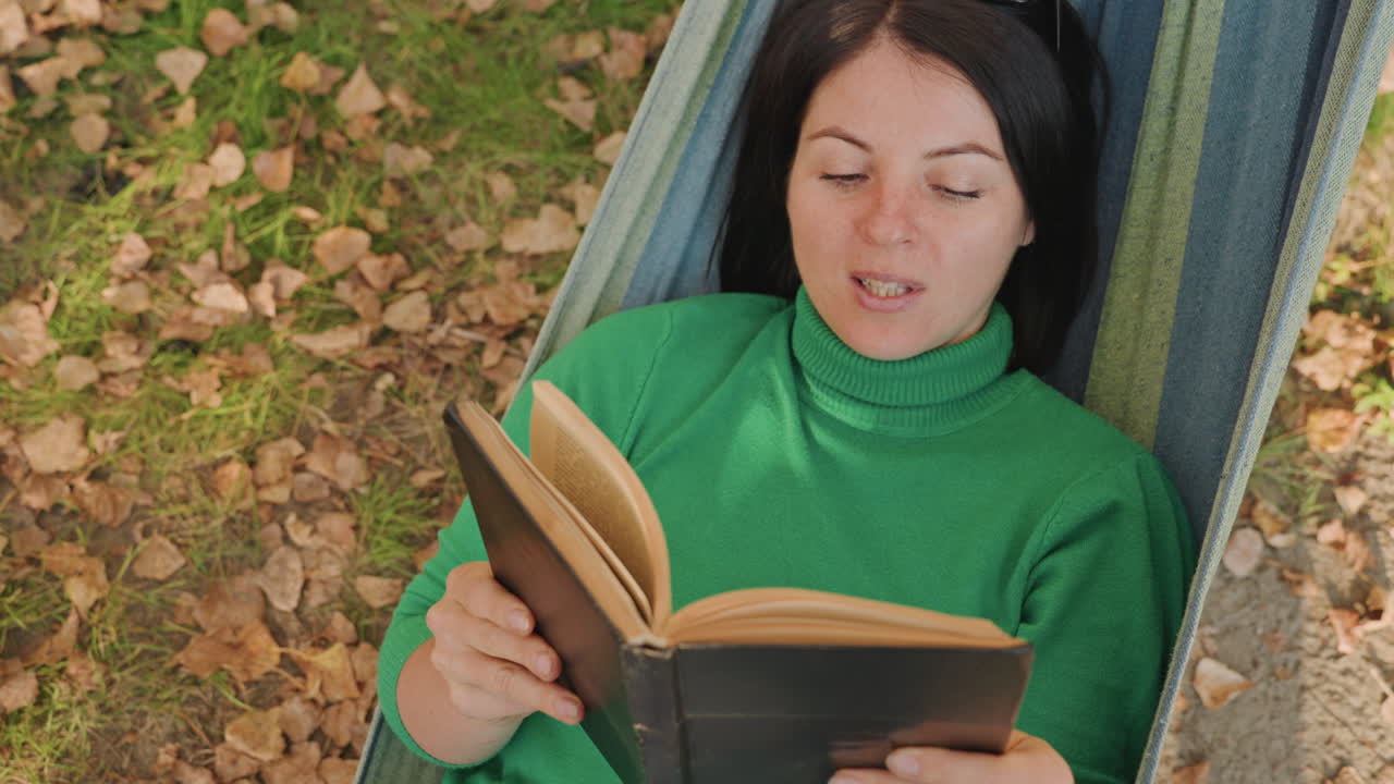 Caucasian woman reading in hammock camping, leaf strewn ground, green sweater, soft autumn light, relaxed expression, hands holding worn book, overhead closeup, meditative outdoor leisure perfect