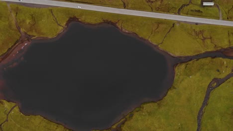 high-up-Aerial-top-down-view-of-huge-lake-with-dark-water-and-small-rivers-in-the-landscape-close-to-a-road-with-cars-driving-by-on-the-Faroe-Islands