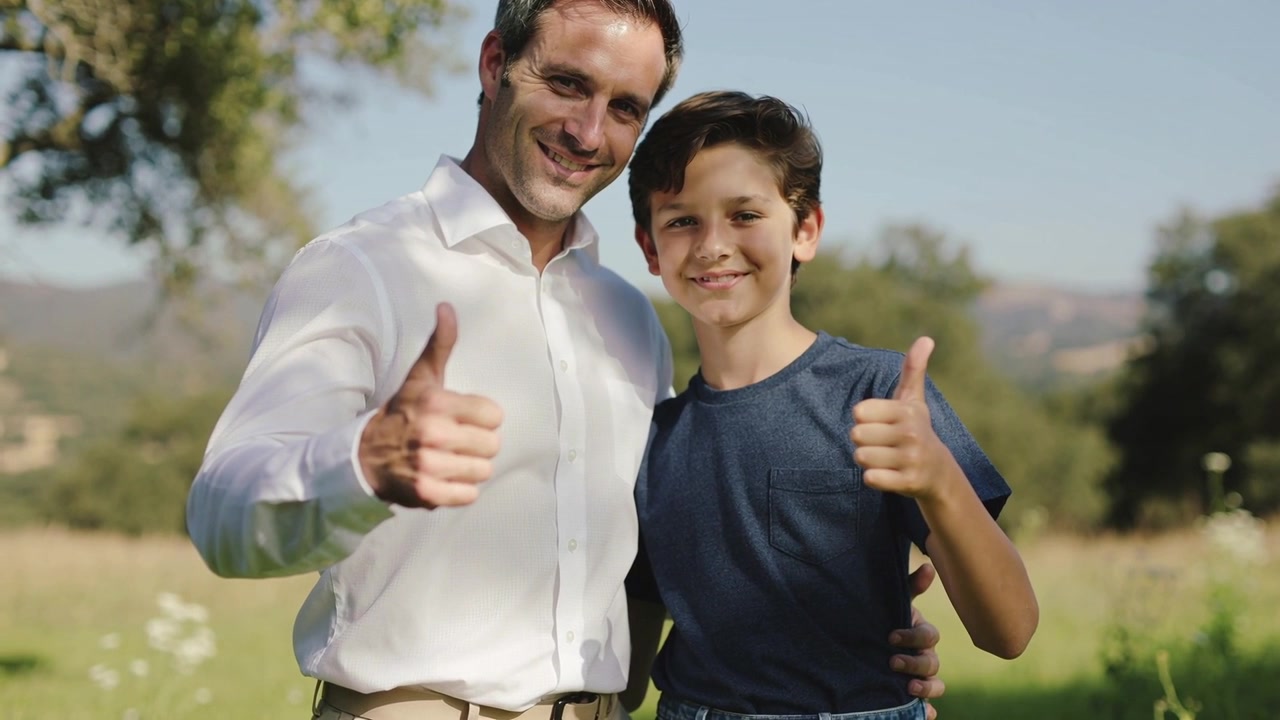 Padre e hijo sonrientes posando al inicio de la toma en un prado, haciendo un gesto de aprobación, con el brazo sobre el hombro, camisa blanca.