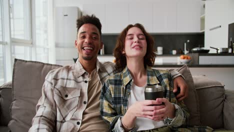 portrait of a happy and very cheerful couple a young brunette man with black skin in a cream plaid shirt sits on the sofa and hugs his girlfriend who is rejoicing and holding a mug of tea in her hands while sitting on a modern sofa in a modern apartment