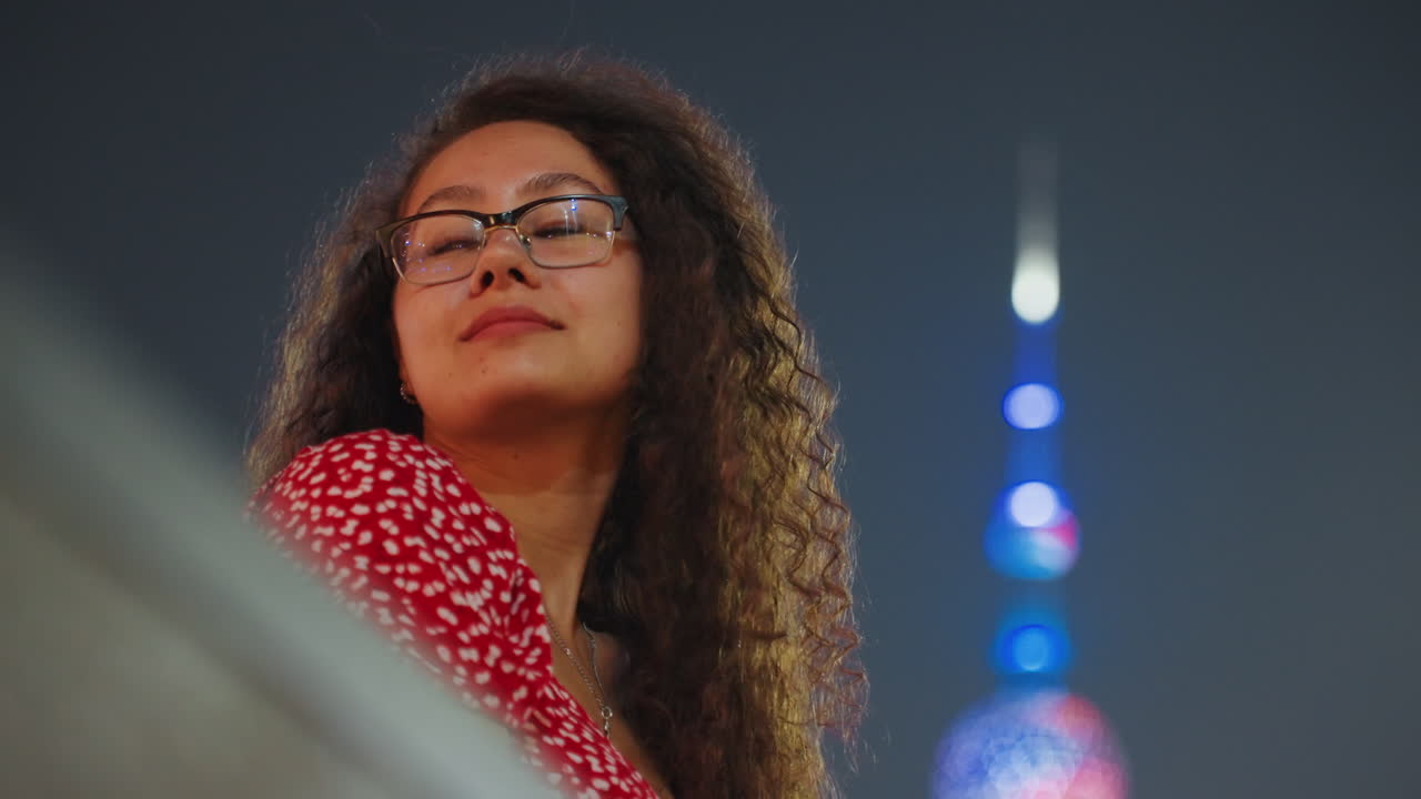 Pensive woman gazing at city lights, curly hair and glasses, red dress silhouette against neon tower, rooftop railing framing, calm reflective mood, cinematic bokeh, quiet night atmosphere