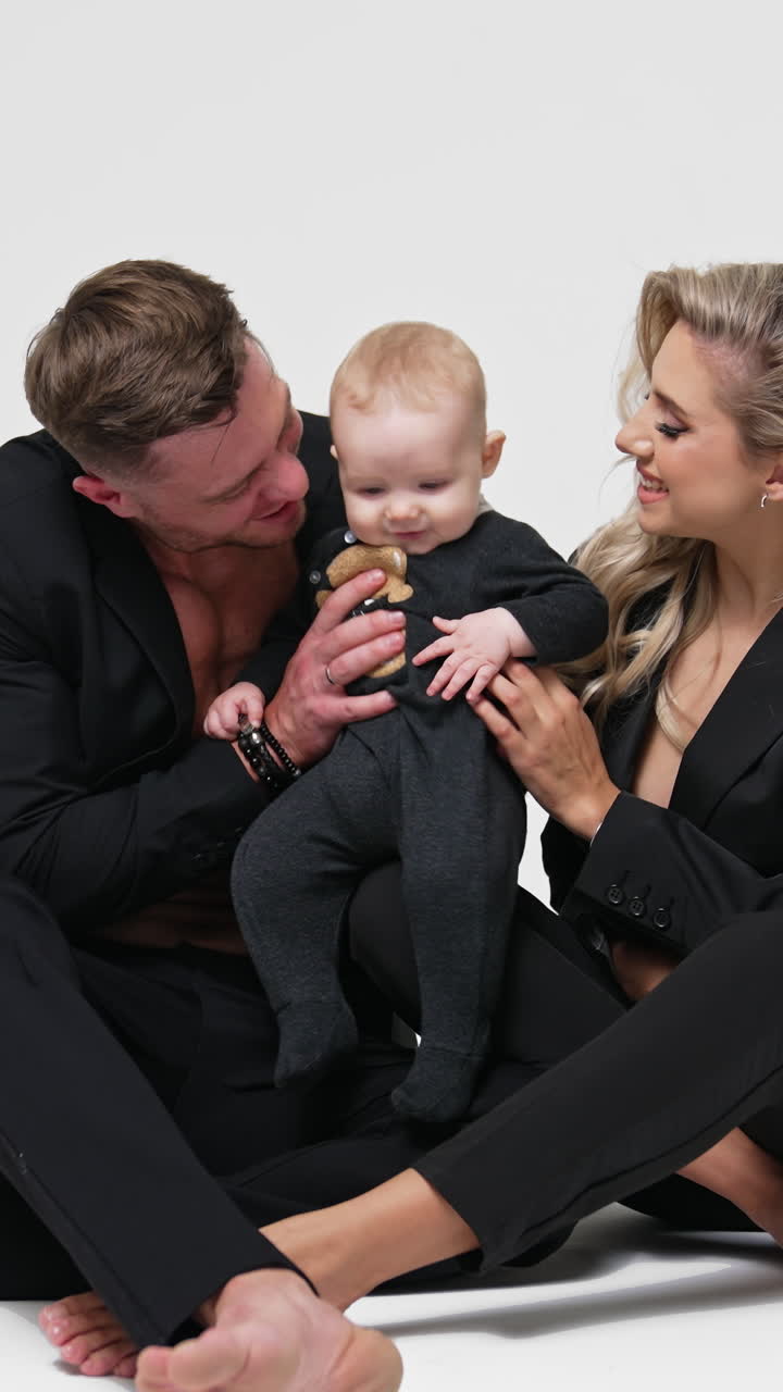 Caucasian couple in black suits sit on the floor with their infant son. Happy young family portrait in studio. White backdrop. Vertical video