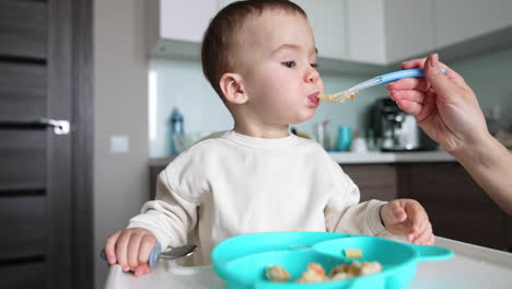 calm adorable baby sits at the feeding table. child waiting for mom giving a full spoon. close up. blurred backdrop.