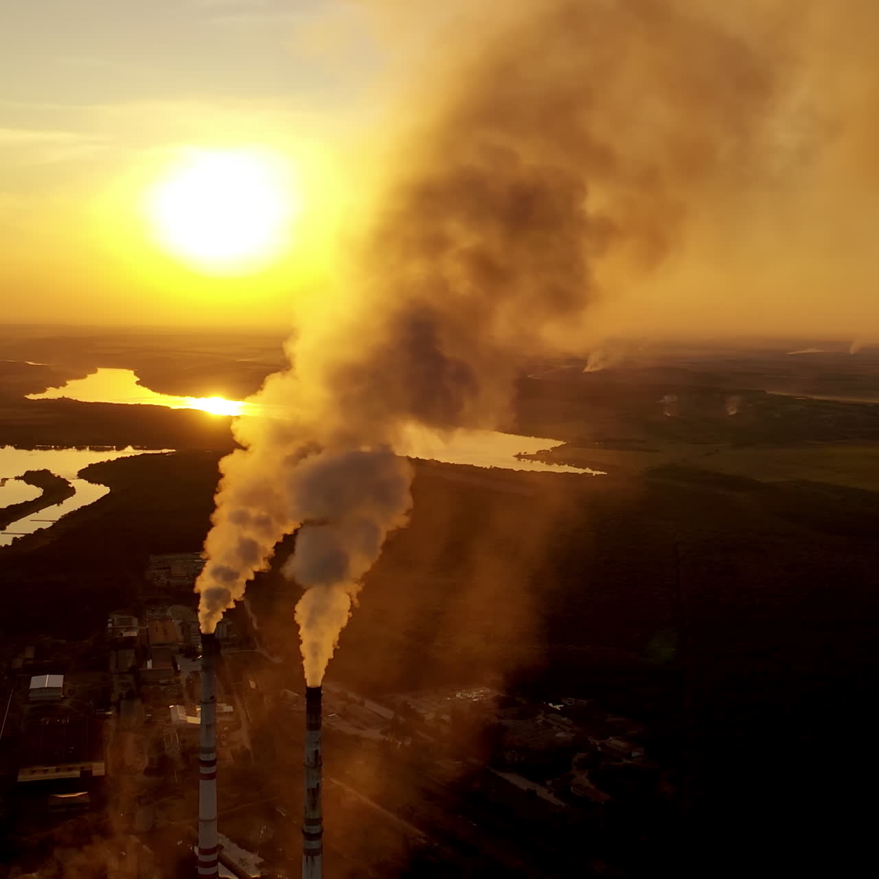 Aerial view on the industrial factory. Power plant near the river on the natural landscape at sunset. Large pipes with smoke in the evening.
