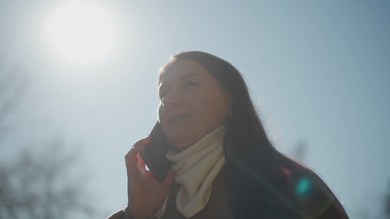 Primo piano di una donna che tiene uno smartphone in mano e passa a parlare al telefono all'aperto sotto la luce solare intensa. La luce del sole crea un effetto lens flare mentre la donna interagisce con il dispositivo contro uno sfondo chiaro