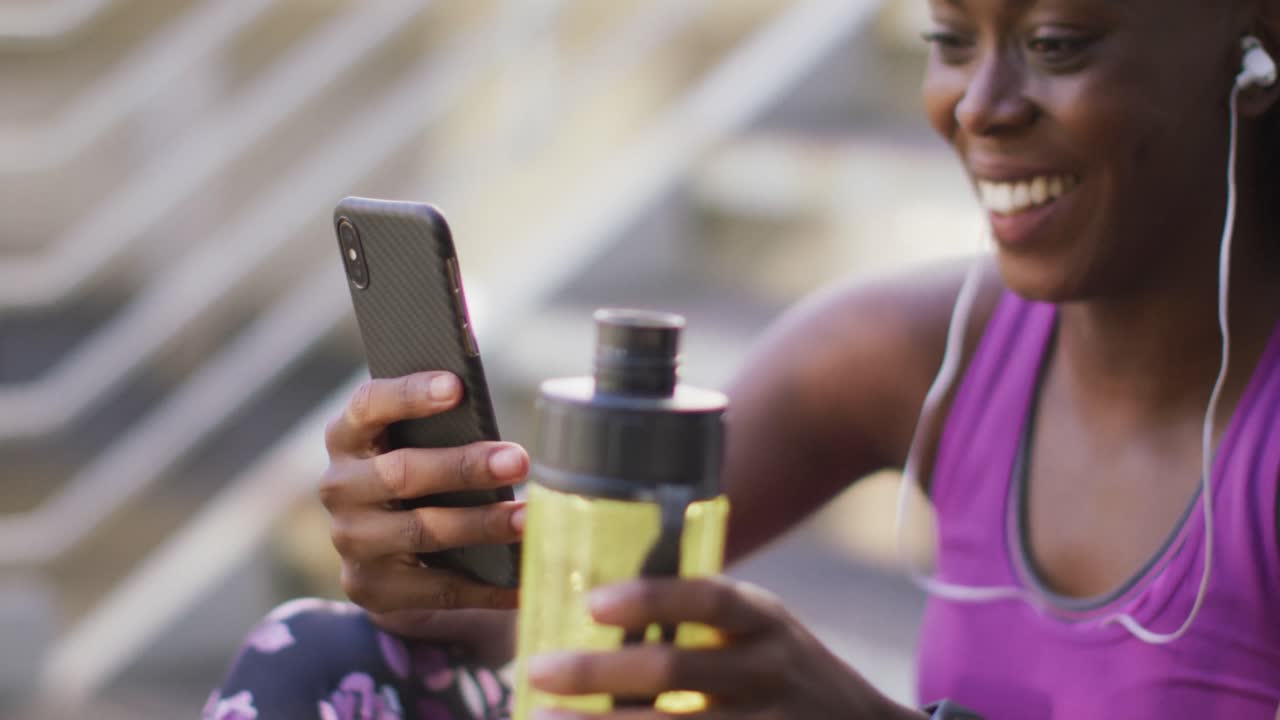 mujer afroamericana sonriendo mientras usa un teléfono inteligente sentada en las escaleras del puente de la ciudad
