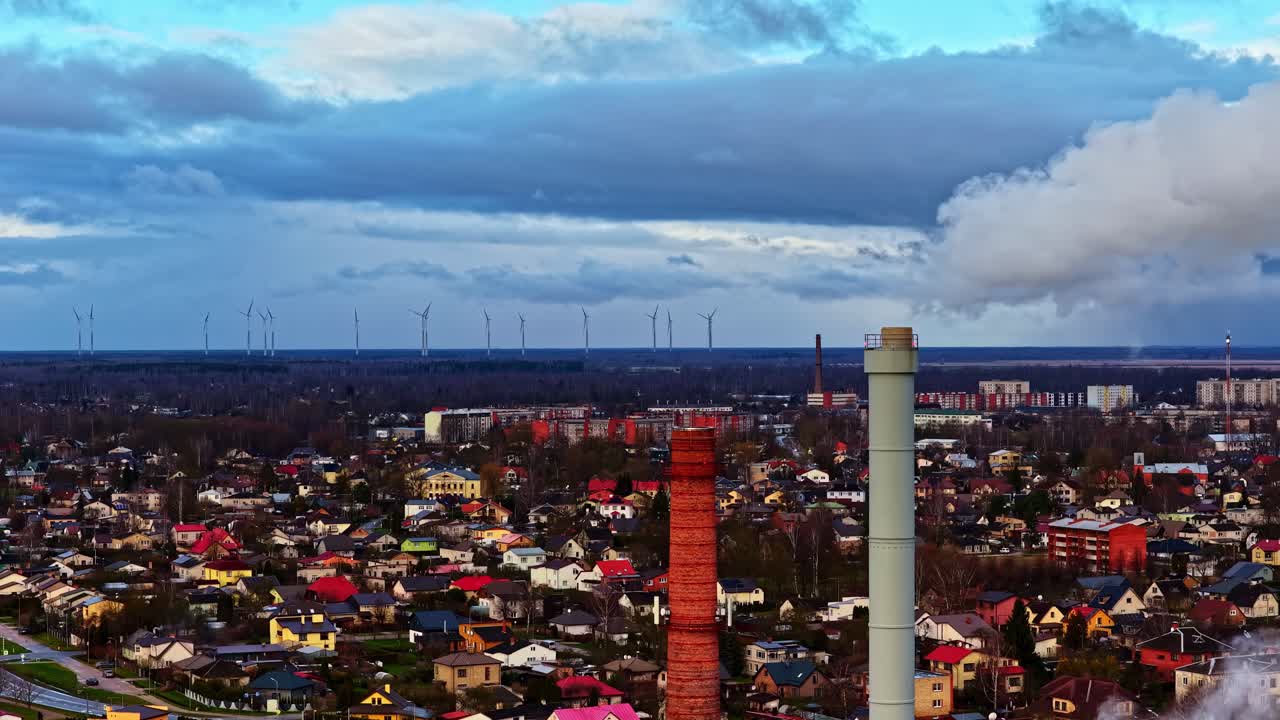 Ripresa aerea con drone di un sobborgo europeo con ciminiere gemelle, pennacchio di scarico e turbine eoliche all'orizzonte sotto drammatiche nuvole temporalesche