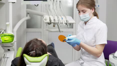 Young-female-dentist-in-mask-and-gloves-using-dental-UV-light-equipment-for-polymer-hardening