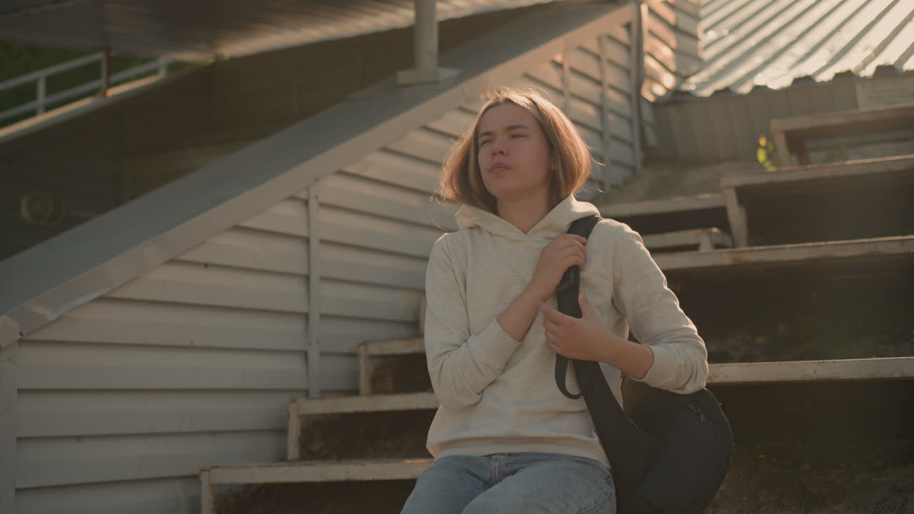 Young woman in casual hoodie holding black bag, standing alone on rustic stadium bleachers, looking down thoughtfully with a worried expression