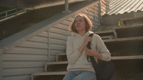 young woman in casual hoodie holding black bag, standing alone on rustic stadium bleachers, looking down thoughtfully with a worried expression